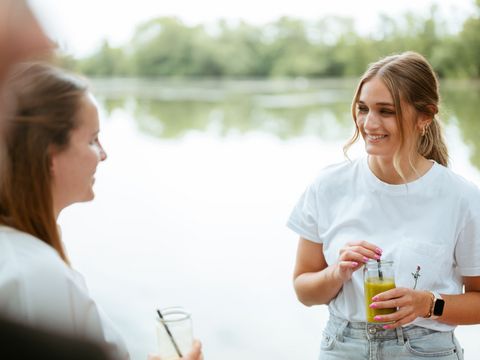 Das Bild zeigt zwei Personen mit Getränken in der Hand. Im Hintergrund ist ein See zu sehen. Die Personen unterhalten sich miteinander. 