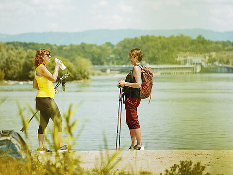 Zwei Wanderer am Seeufer Zwei Damen mit Wanderstöcken und Freizeitkleidung machen eine Pause vor dem Rhein, im Hintergrund das Wasserkraftwerk von Rheinfelden.