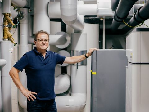 Das Bild zeigt eine Person neben einer Wärmepumpe, einem Gerät zur effizienten Heizung und Kühlung. Wärmepumpen funktionieren, indem sie Wärme von einem Ort (z.B. Außenluft oder Erde) aufnehmen und in einen anderen übertragen, um Gebäude zu heizen oder zu kühlen. Sie gelten als umweltfreundliche und energieeffiziente Alternative zu herkömmlichen Heizsystemen, da sie weniger fossile Brennstoffe verbrauchen. In vielen modernen Häusern werden sie aufgrund ihrer Fähigkeit, Energie zu sparen, zunehmend eingesetzt.