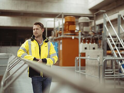 Ein Mann in einer gelben Sicherheitsjacke steht in einer industriellen Umgebung. Im Hintergrund sind verschiedene Maschinen und Metallstrukturen zu sehen.